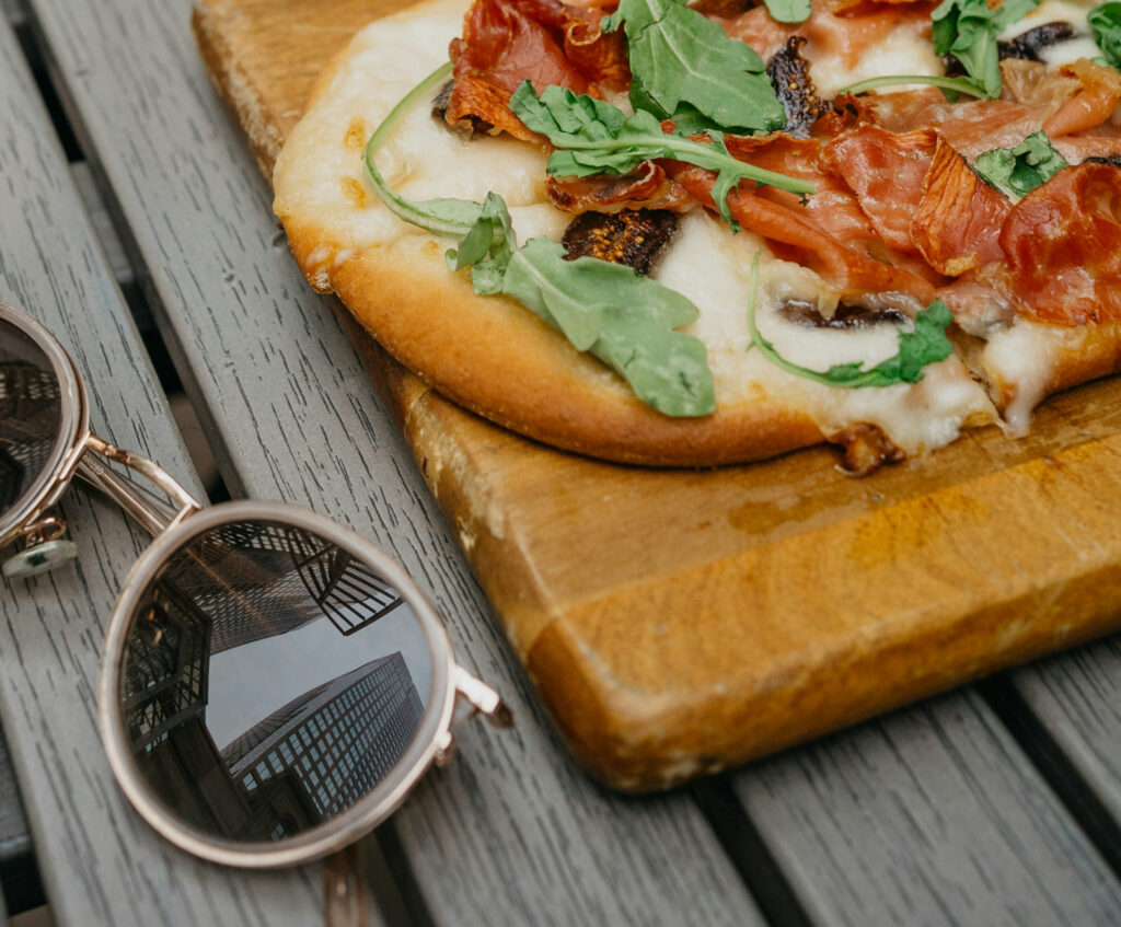 Flat bread on wood board with prosciutto and arugula on picnic table with sunglasses