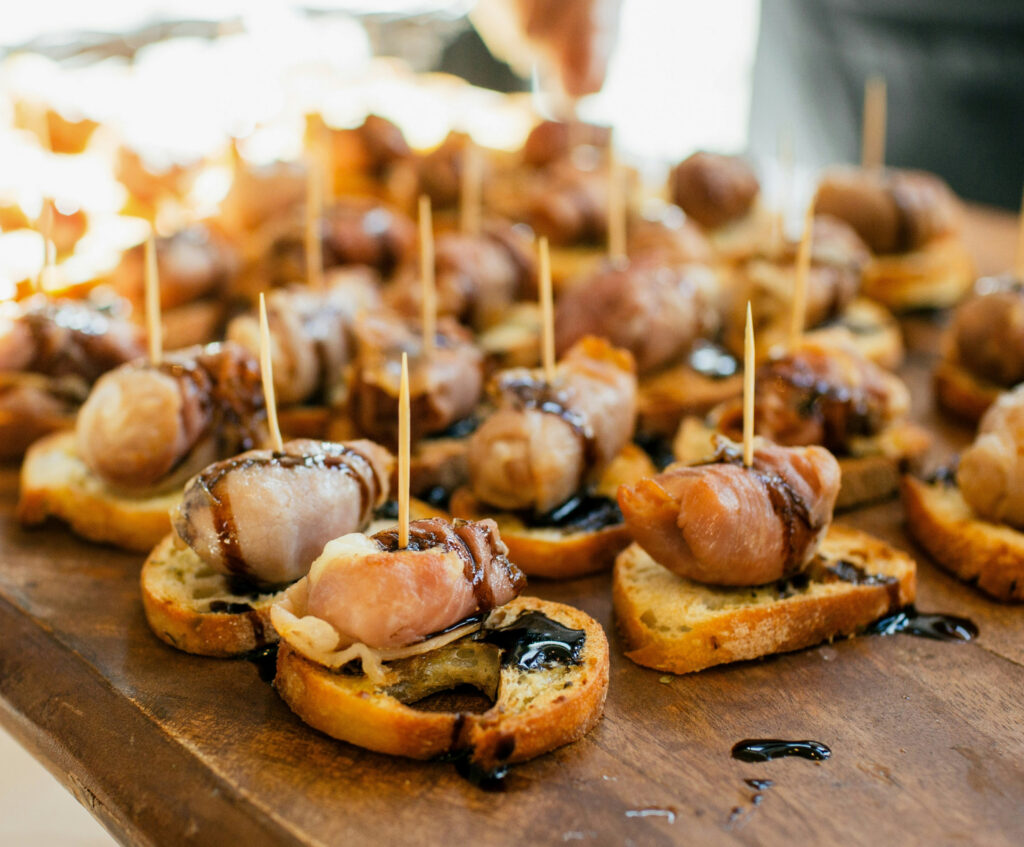 Close up of many crostini with fancy meat and sauce drizzle, speared with toothpicks, on wood board