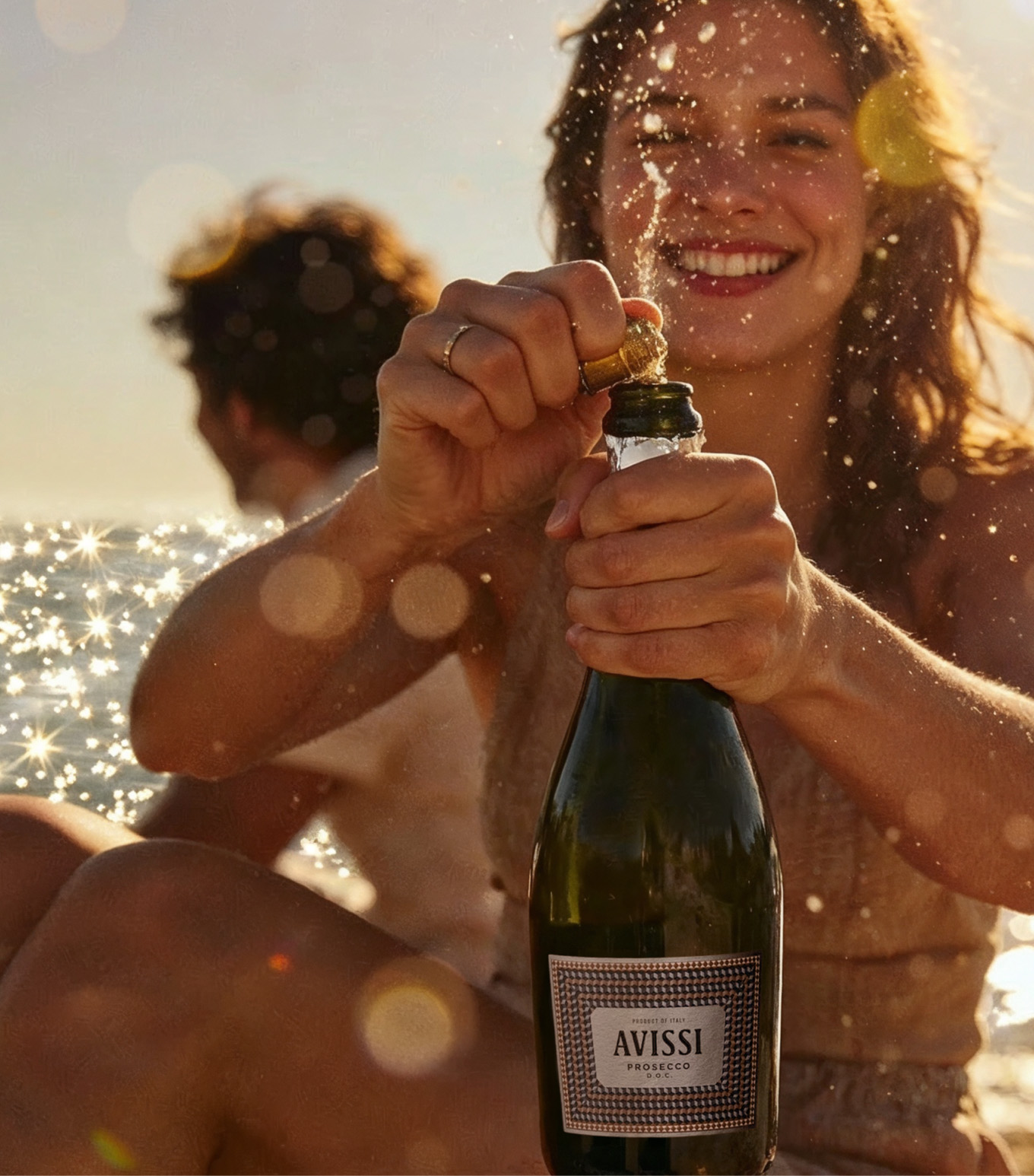 A tanned woman opening a bottle of Avissi Prosecco in front of her with a man behind her turned away and sparking ocean in the background
