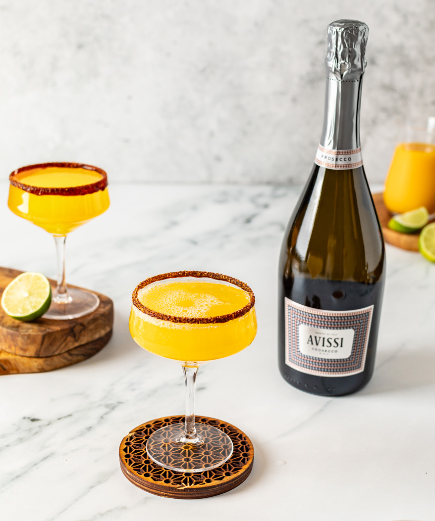 A table top arranement of two prosecco cocktails and a bottle of Avissi Prosecco, with a bottle of orange juice and limes in the background