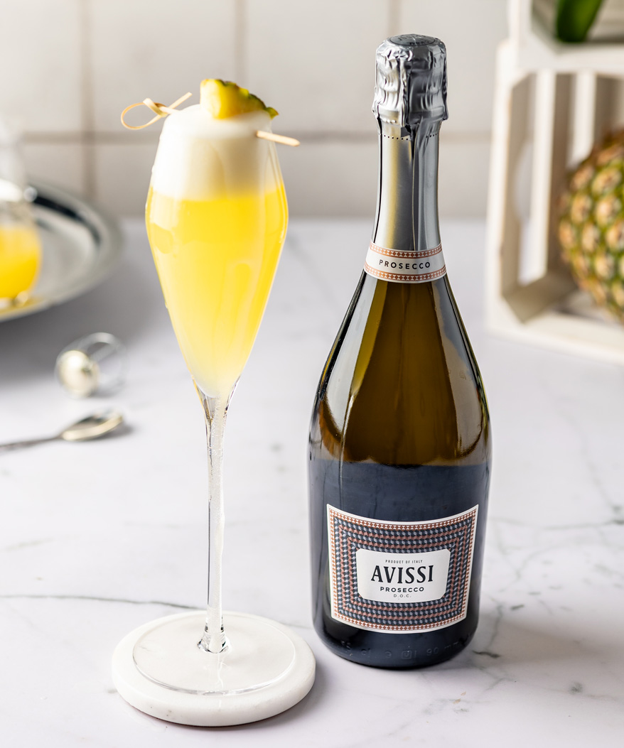 A table top arrangement of a mimosa cocktail and a bottle of Avissi Prosecco, with various bits of fruit and cocktail making tools in the background