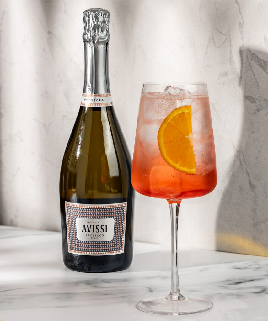A table top arrangement of an aperol spritz and a bottle of Avissi Prosecco