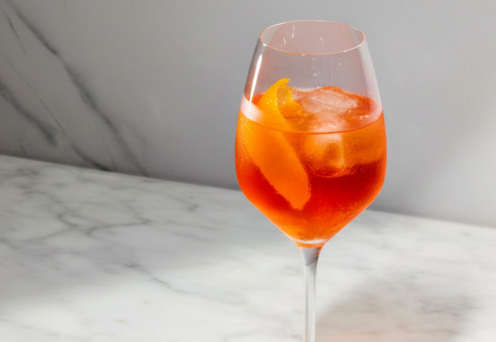 Close up of an orange-red cocktail in a wine glass on a marble surface