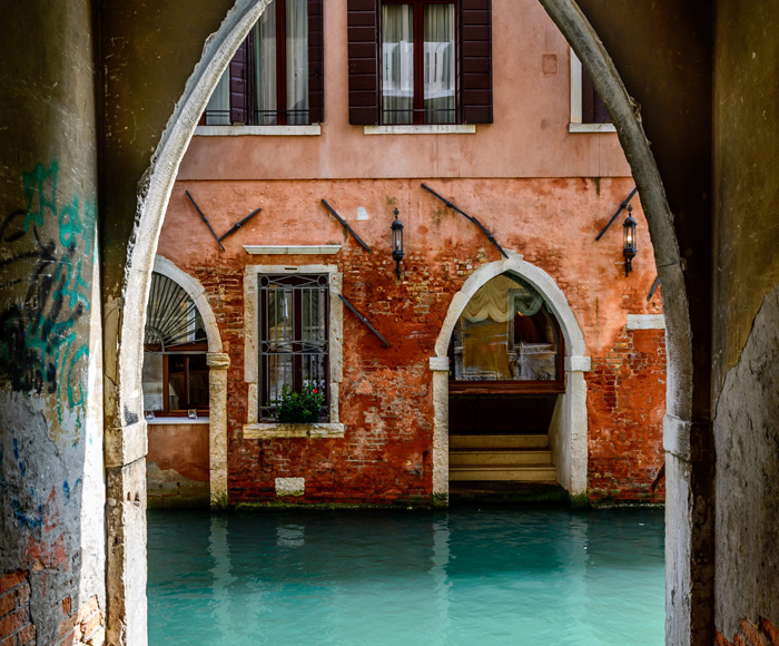 A venice scene showing a two story stucco building with arched doorways and exposed brick with a canal as seen through an arched doorway with graffiti on its walls