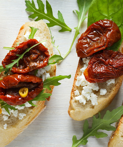 A close up of two toasted crustini with arugula, crumbled cheese, and sun dried tomatoes