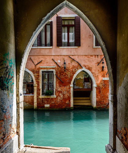A venice scene showing a two story stucco building with arched doorways and exposed brick with a canal as seen through an arched doorway with graffiti on its walls